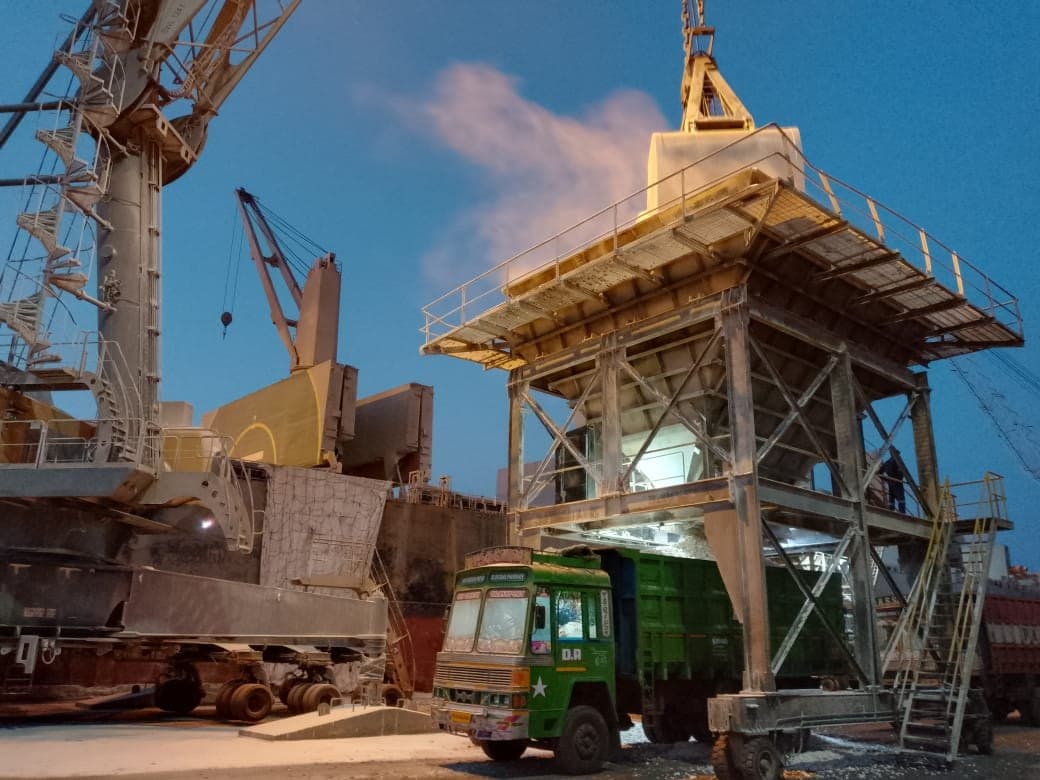 Grab crane feeding a dockside hopper loading a tipper truck beside a vessel at night