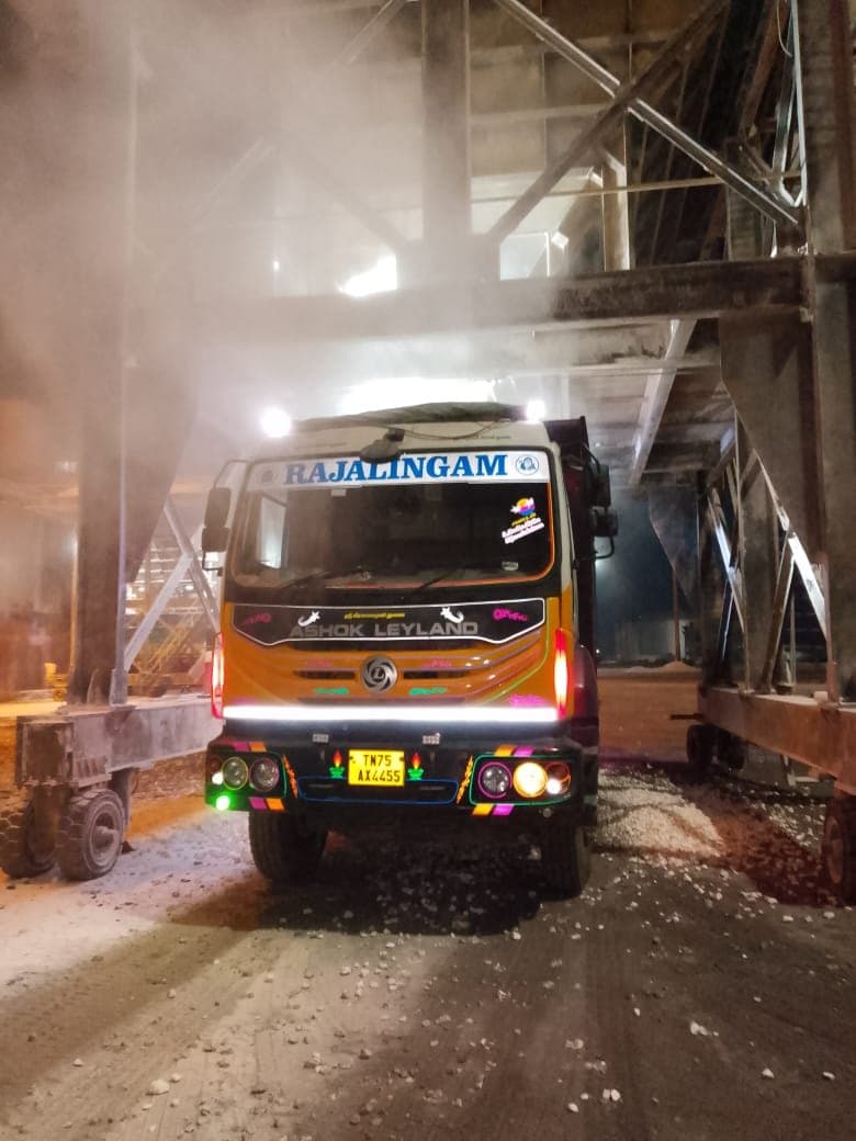 Tipper truck under a loading hopper with dust suppression spray at a night-time port yard