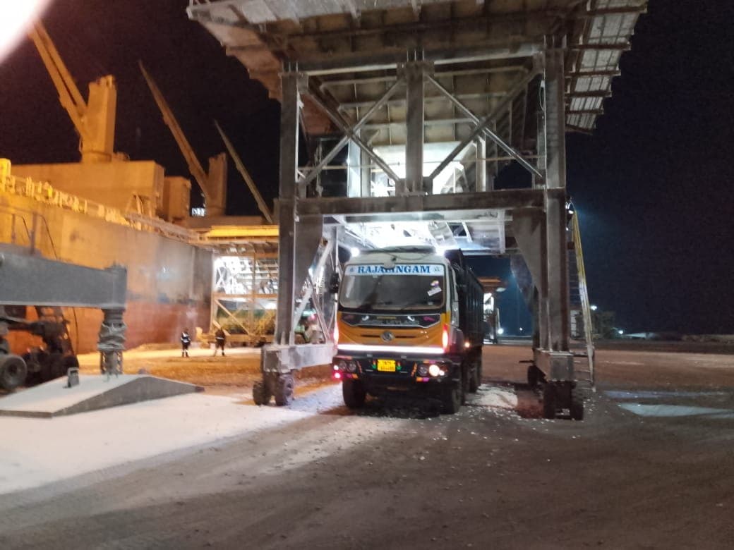 Truck positioned beneath a mobile hopper next to a docked vessel during night cargo evacuation