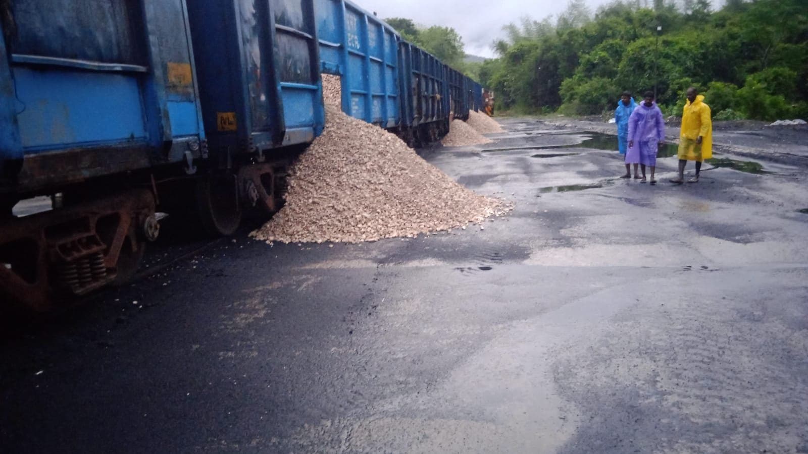 Rake of wagons unloading crushed stone beside workers in raincoats on a wet yard