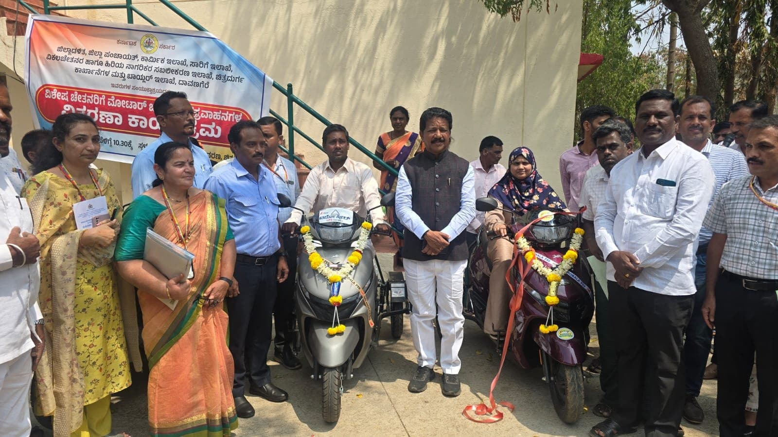 Beneficiaries seated on garlanded scooters flanked by officials at a handover ceremony